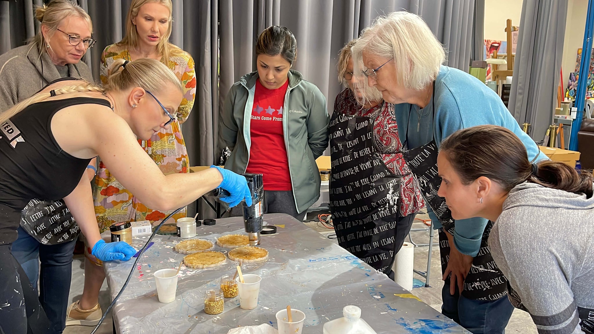 Anja Yamaji uses a heat gun to create gold resin coasters while workshop participants gather around to watch.