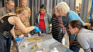 Anja Yamaji uses a heat gun to create gold resin coasters while workshop participants gather around to watch.