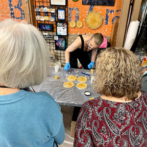 Anja Yamaji demonstrates how to use a toothpick to create a design in resin coasters as students watch her attentively.